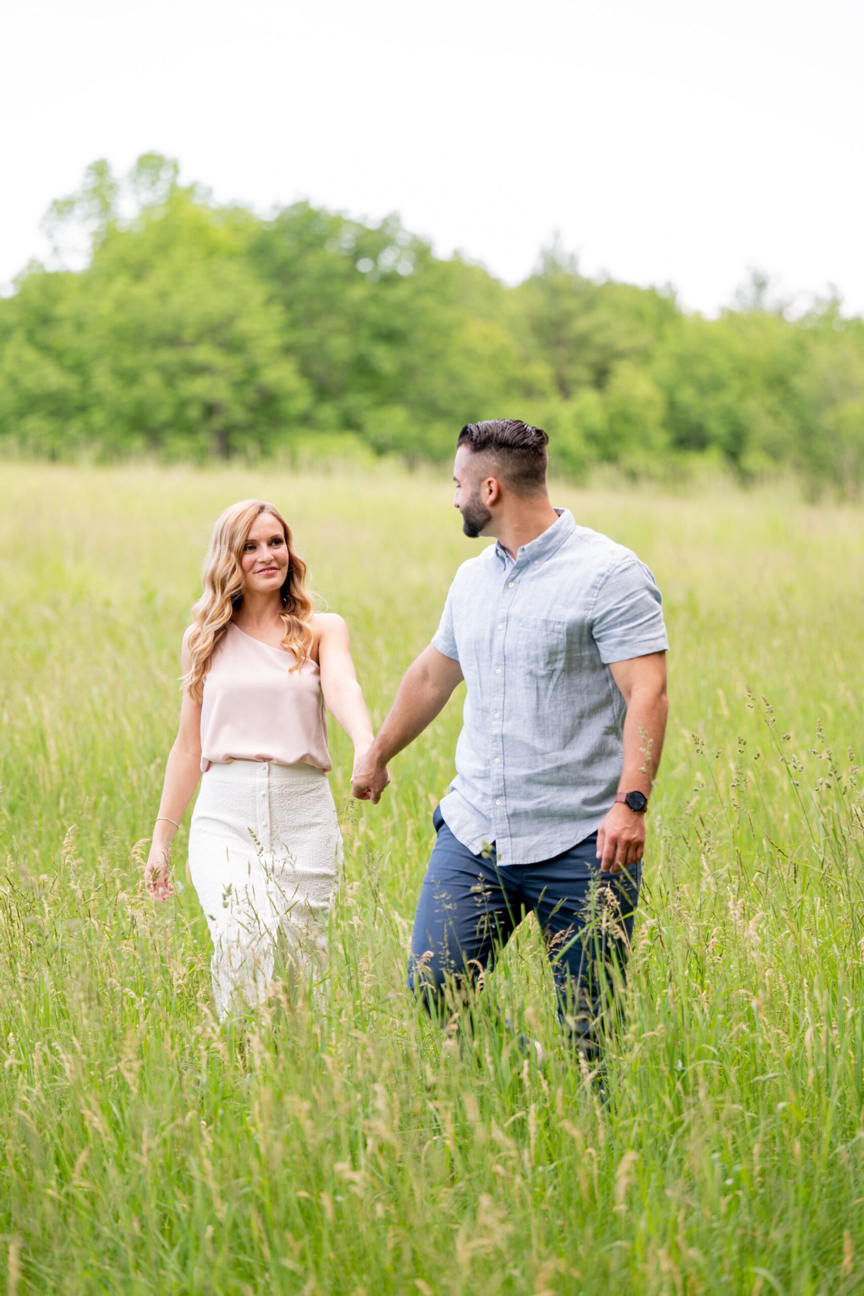 couple holding hands and walking through a green grassy field with green trees behind them