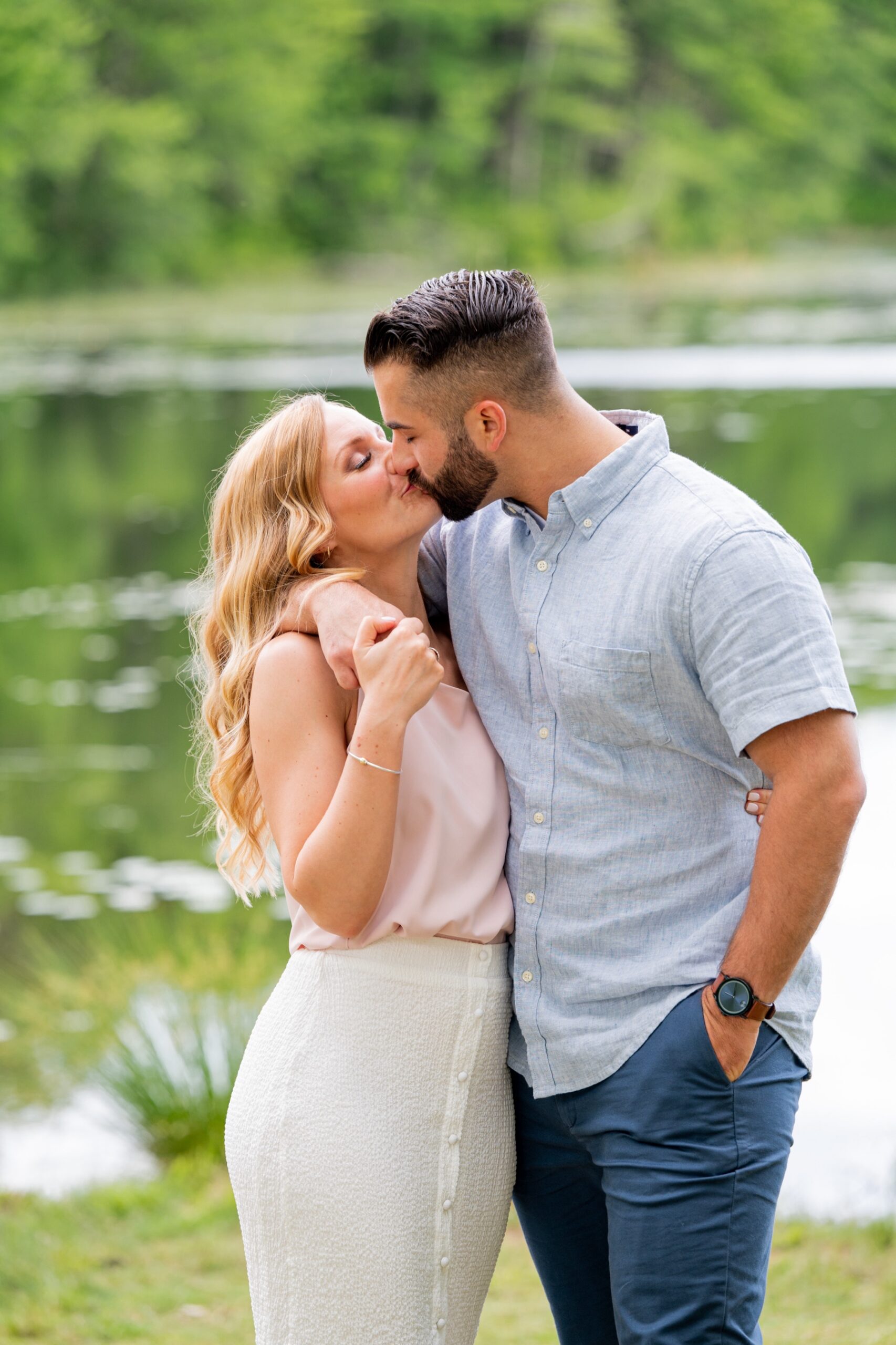 couple kissing in front of a lake with greenery surrounding them