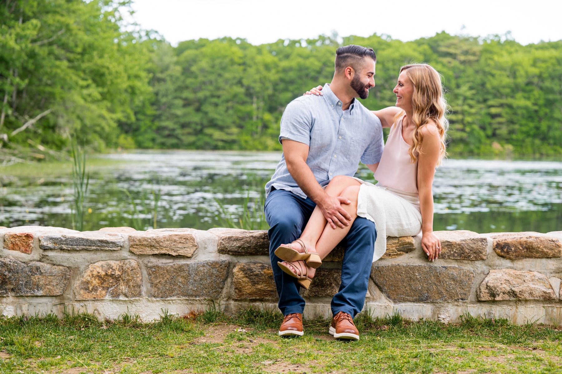 man wearing blue jeans and blue button down shirt and woman wearing pink and white dress sit on stone wall with pond behind them