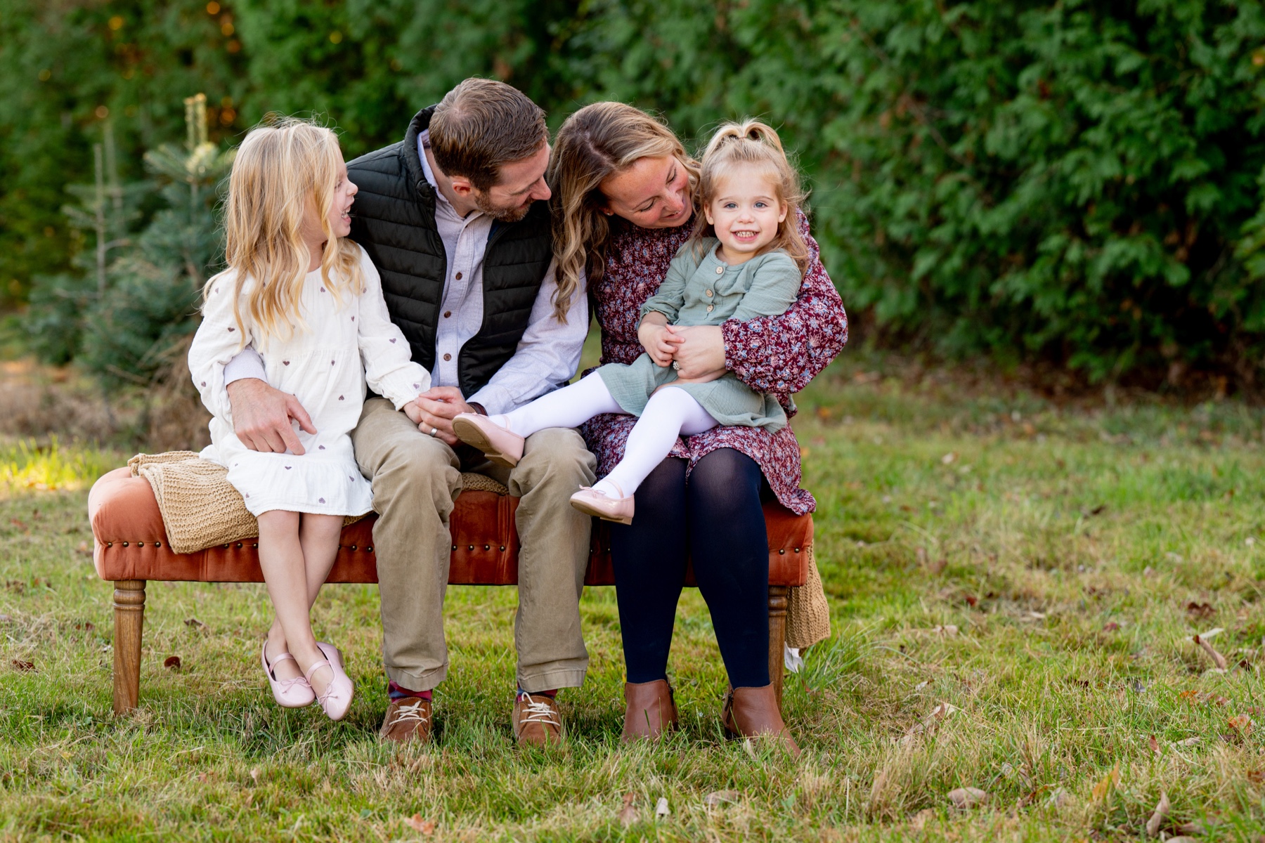 mother and father sitting on a bench with their two daughters surrounded by green trees 
