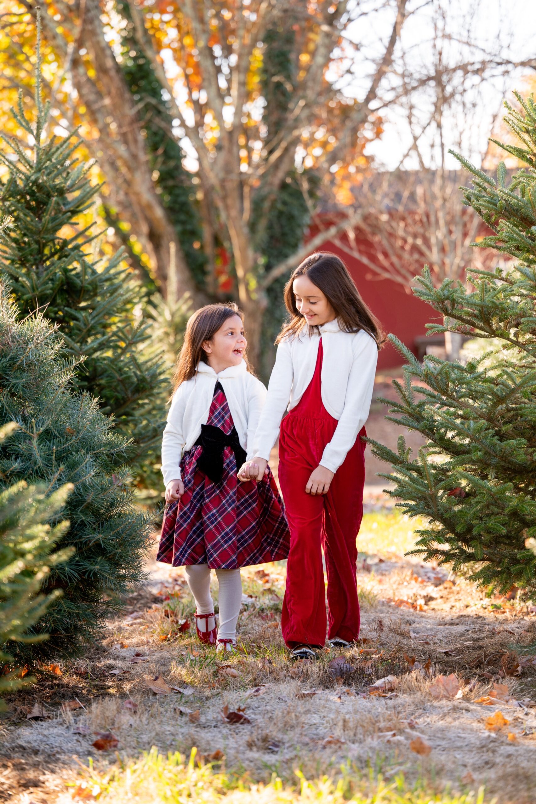 two young girls wearing red outfits walking through a row of christmas trees