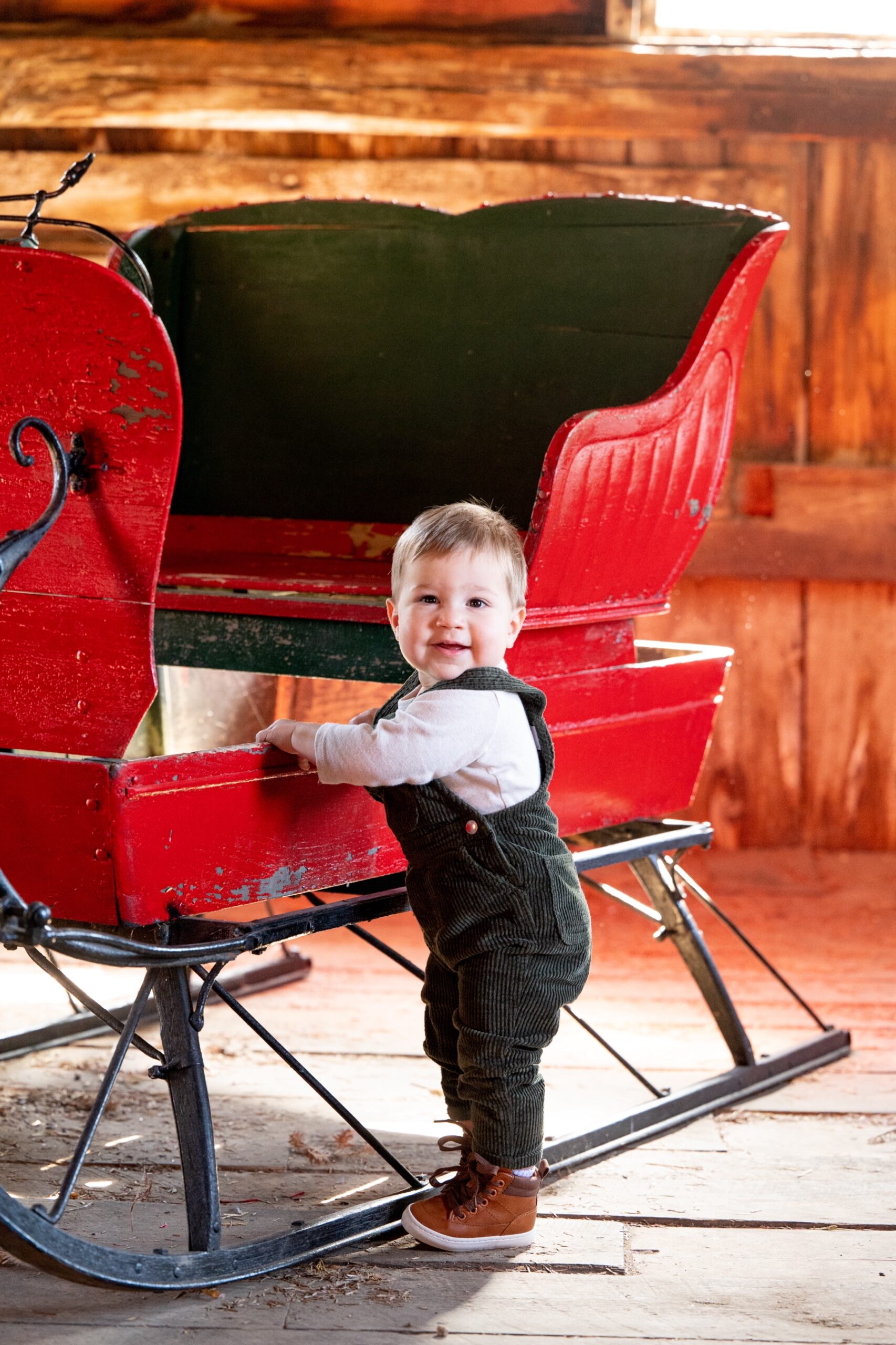 young boy wearing overalls standing next to red holiday sleigh 