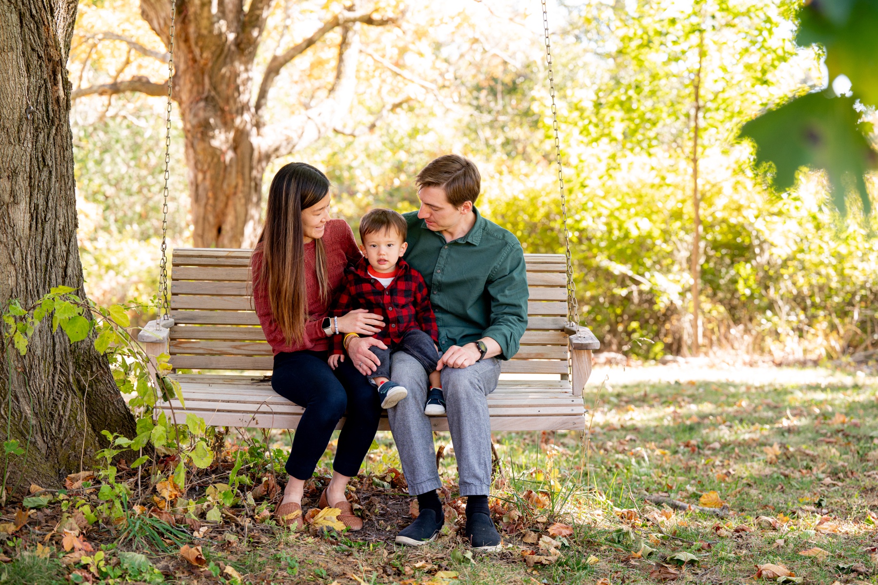 parents sitting with young boy sitting on wooden swing bench surrounded by fall foliage 