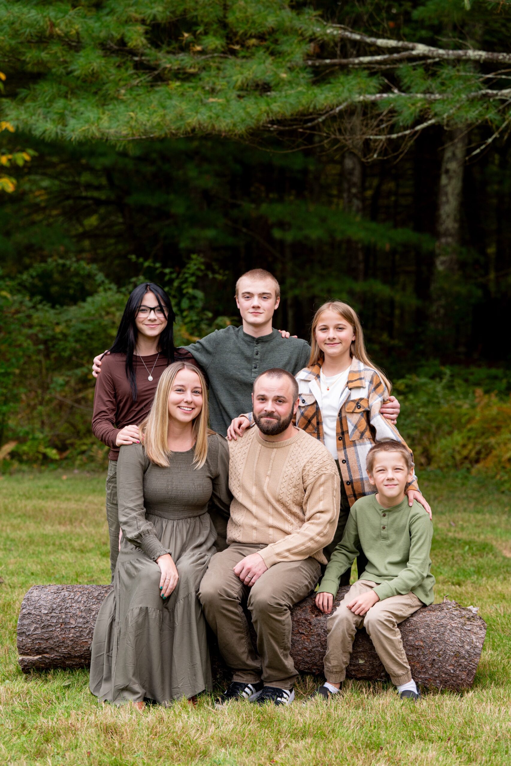 family wearing green and tan and brown outfits sits on a log in sterling ma portrait session