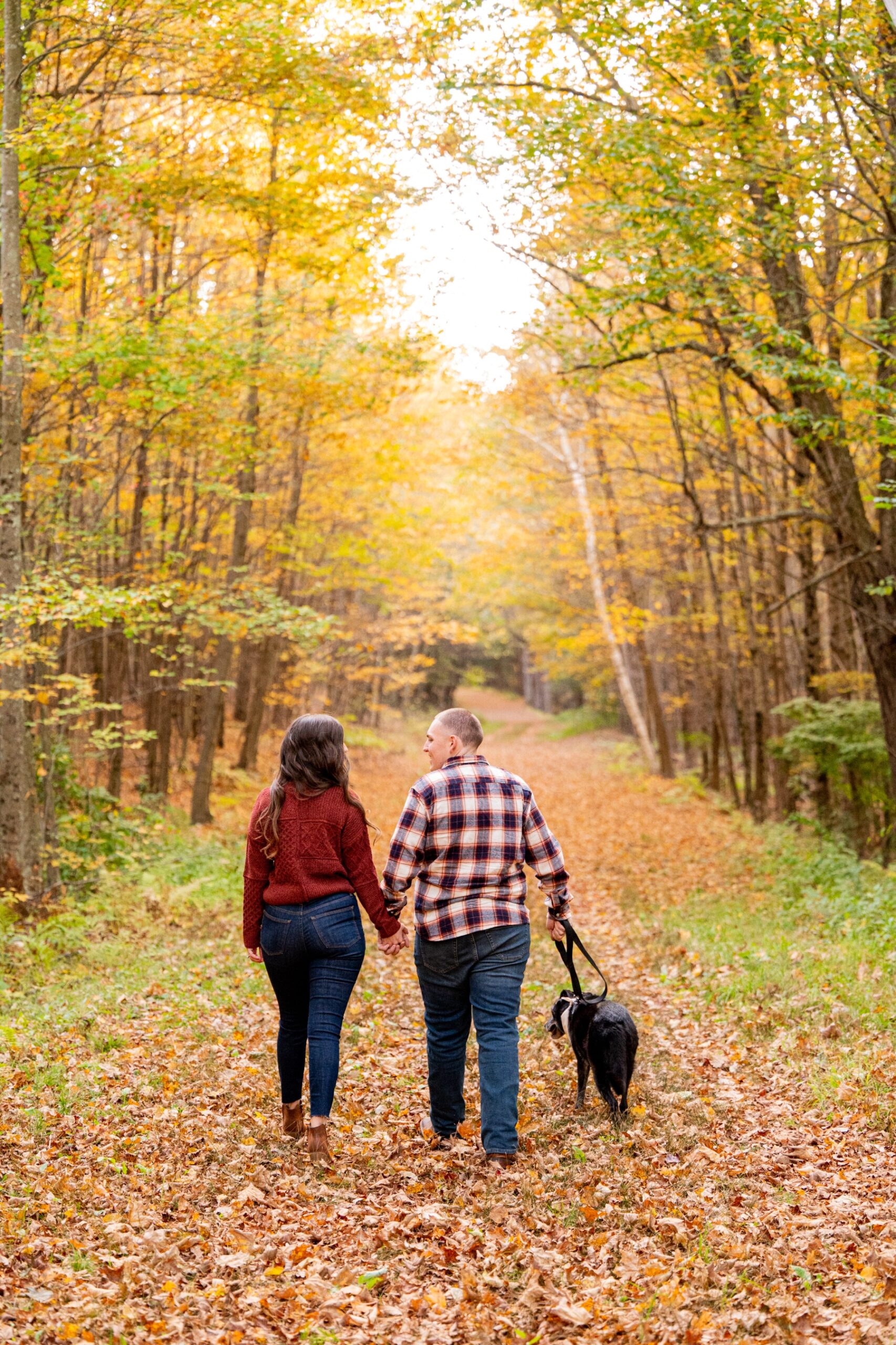 man and woman walking dog through the woods with bright orange and yellow leaves for engagement session