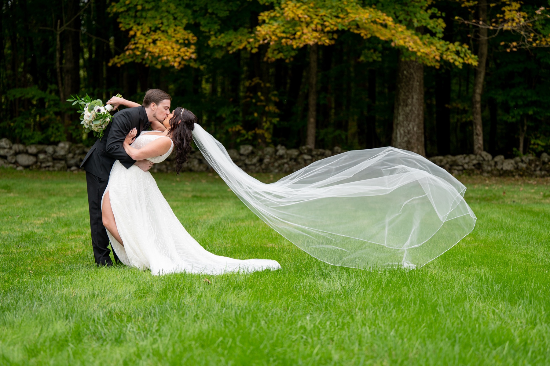 bride and groom kissing in green grassy backyard with veil flowing in the wind