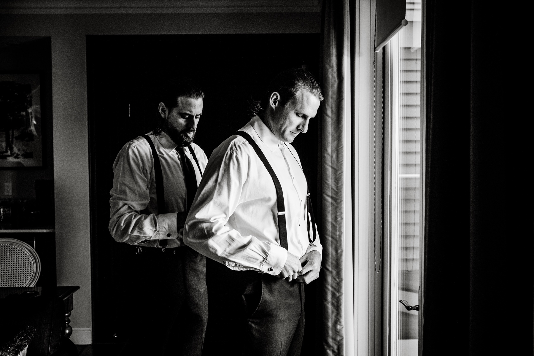 groom standing in front of a window as he puts on his suspenders 