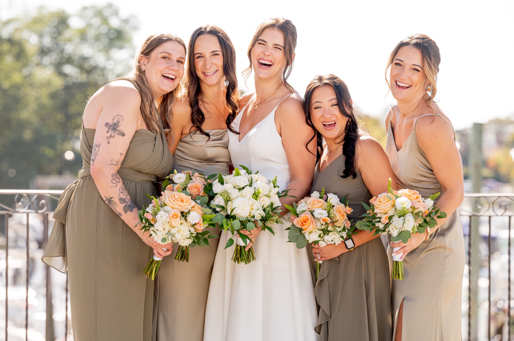 bride with her bridesmaids wearing sage green and light tan dresses with white and orange flowers