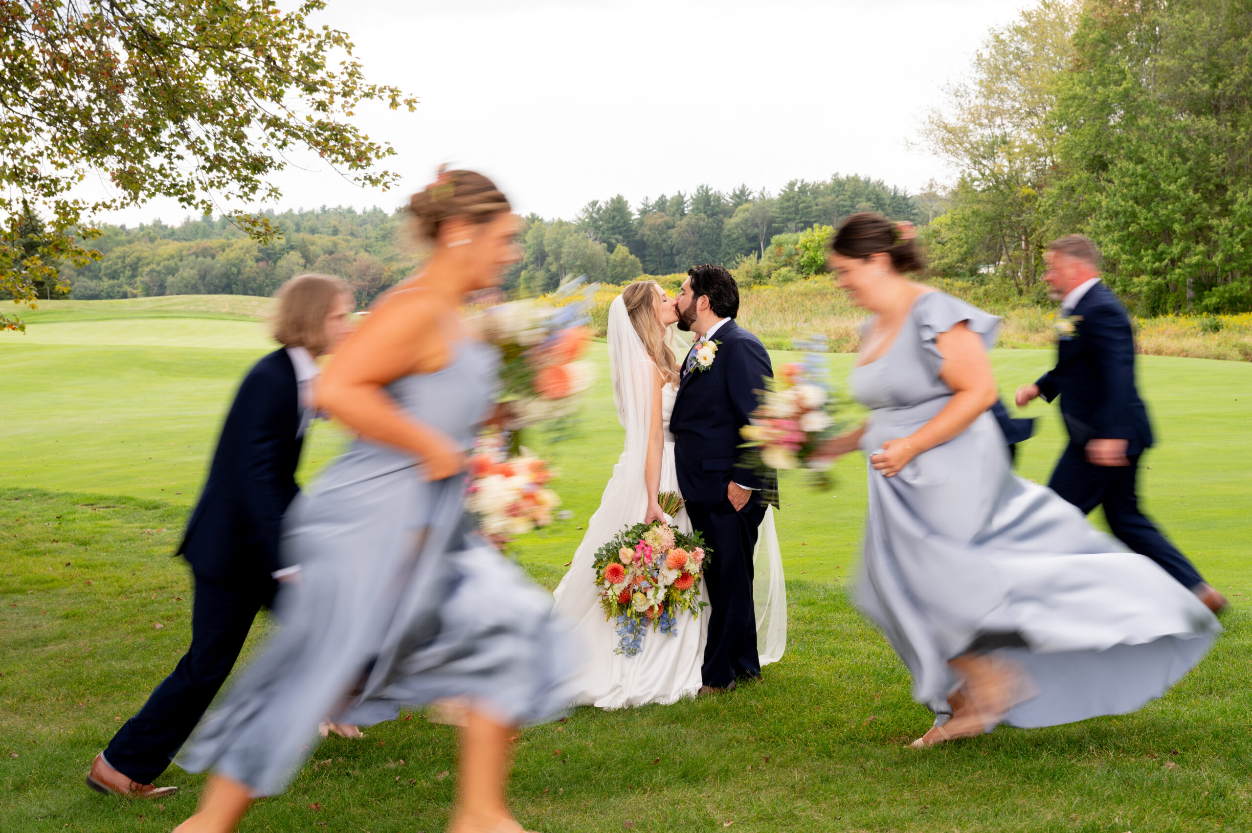 Bride and groom kiss while bridal party runs past them in a blur - exactly what a wedding content creator in New England would capture
