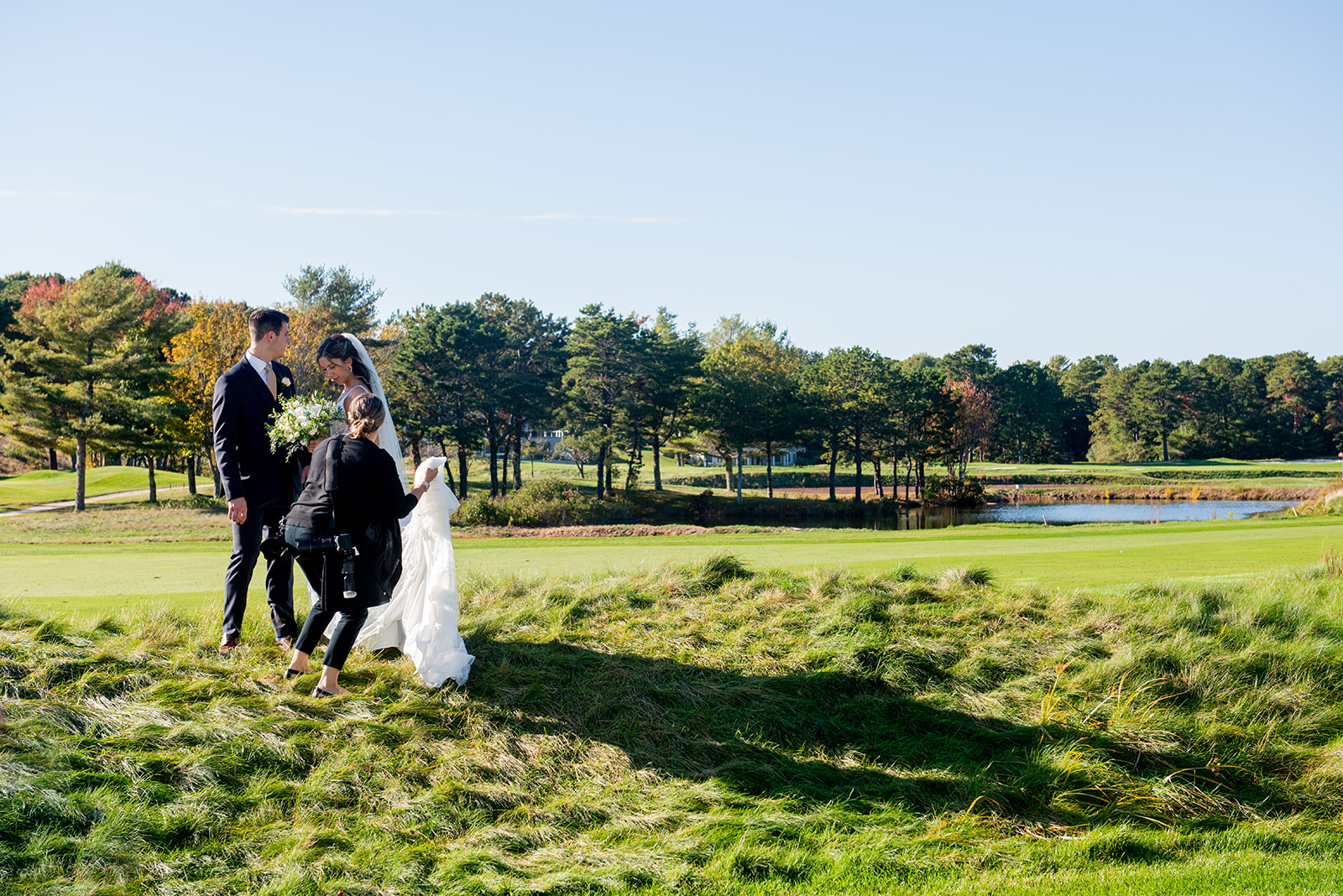 wedding photographer adjusting bride's dress for photos on grassy lawn