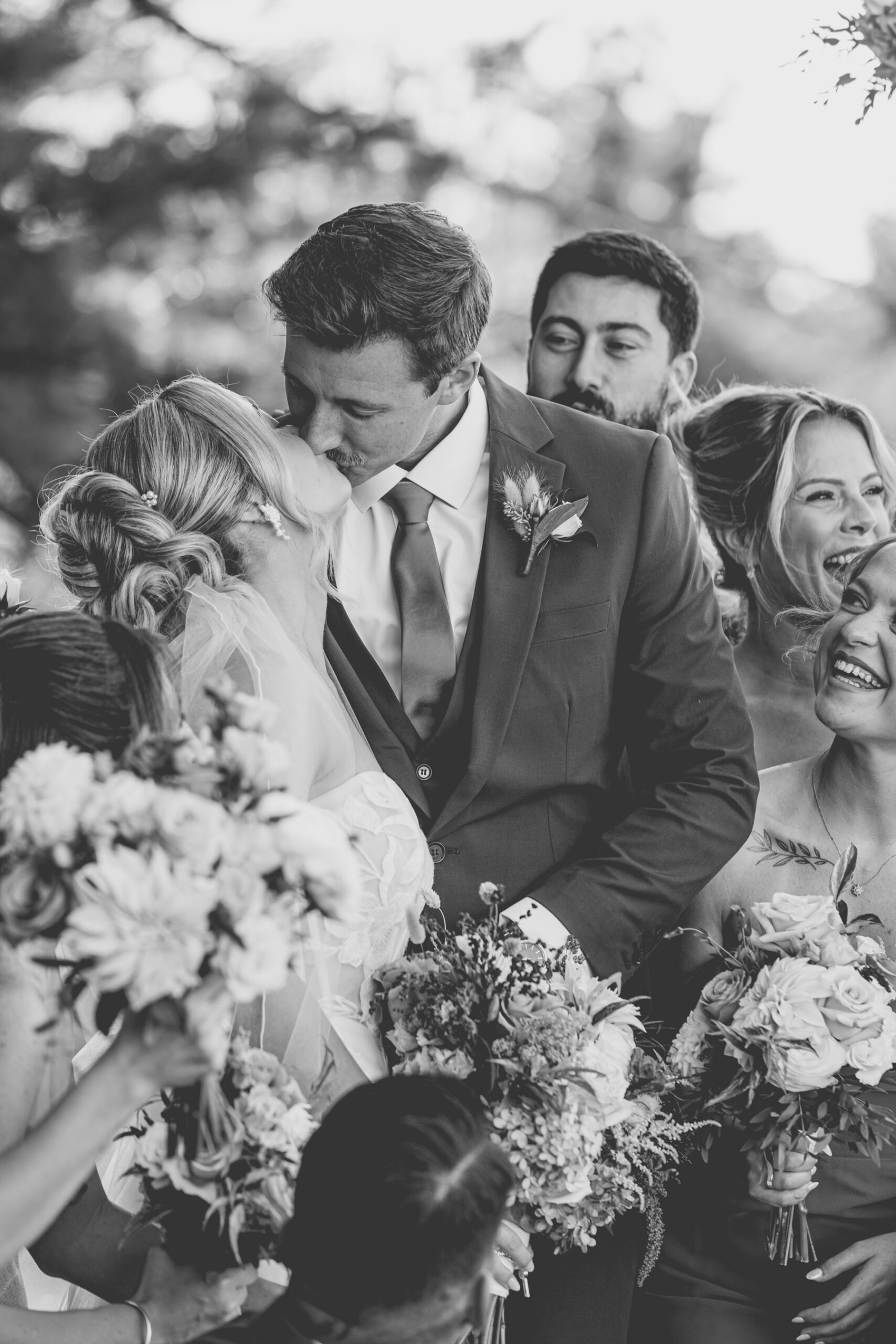 Black and white close up photo of bride and groom kissing surrounded by bridal party
