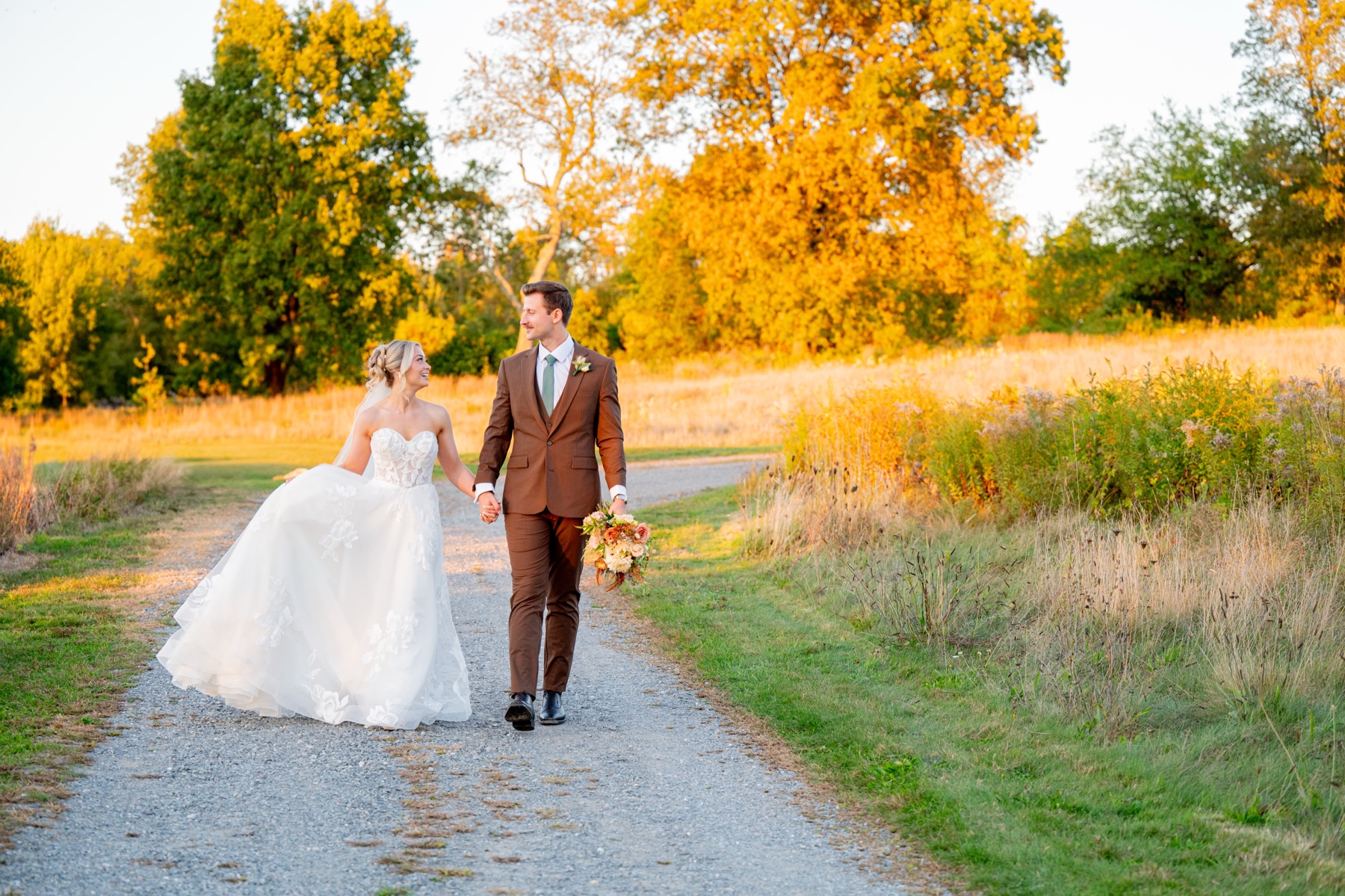 Bride and groom walking on gravel road surrounded by trees with evening sun filtering in the background