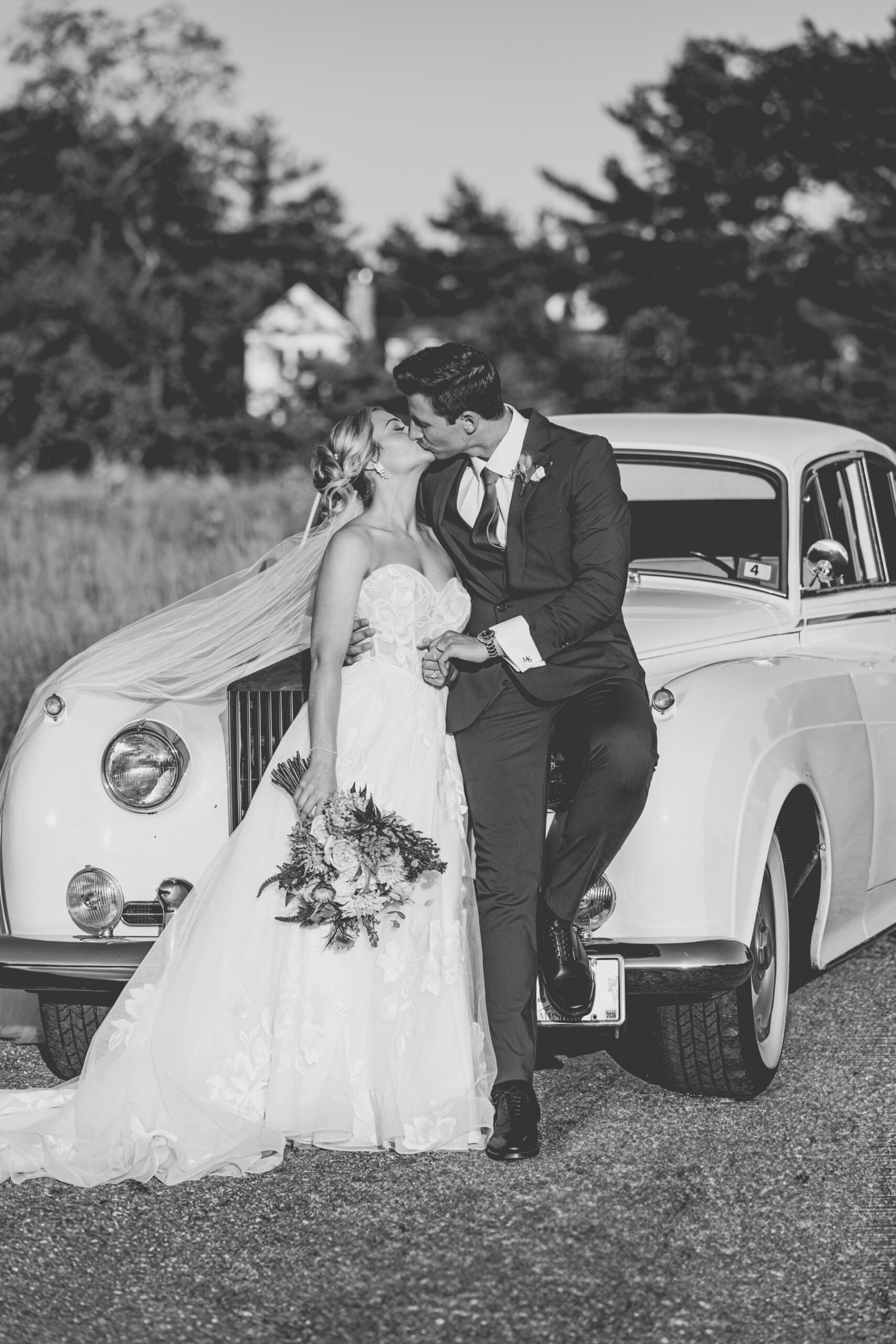 Black and white photo of bride and groom kissing while standing in front of white vitage car